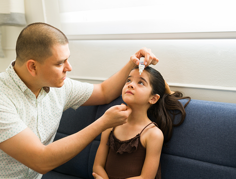 Father administering allergy eye drops to his young daughter while she sits on a couch and looks up